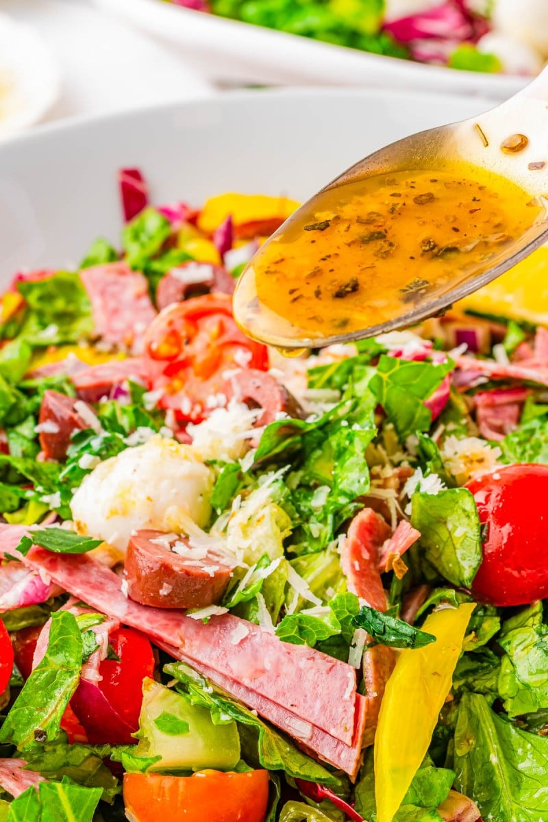 A close-up of an Italian salad with sliced meats, cherry tomatoes, cheese, and greens, as dressing is being poured from a spoon.