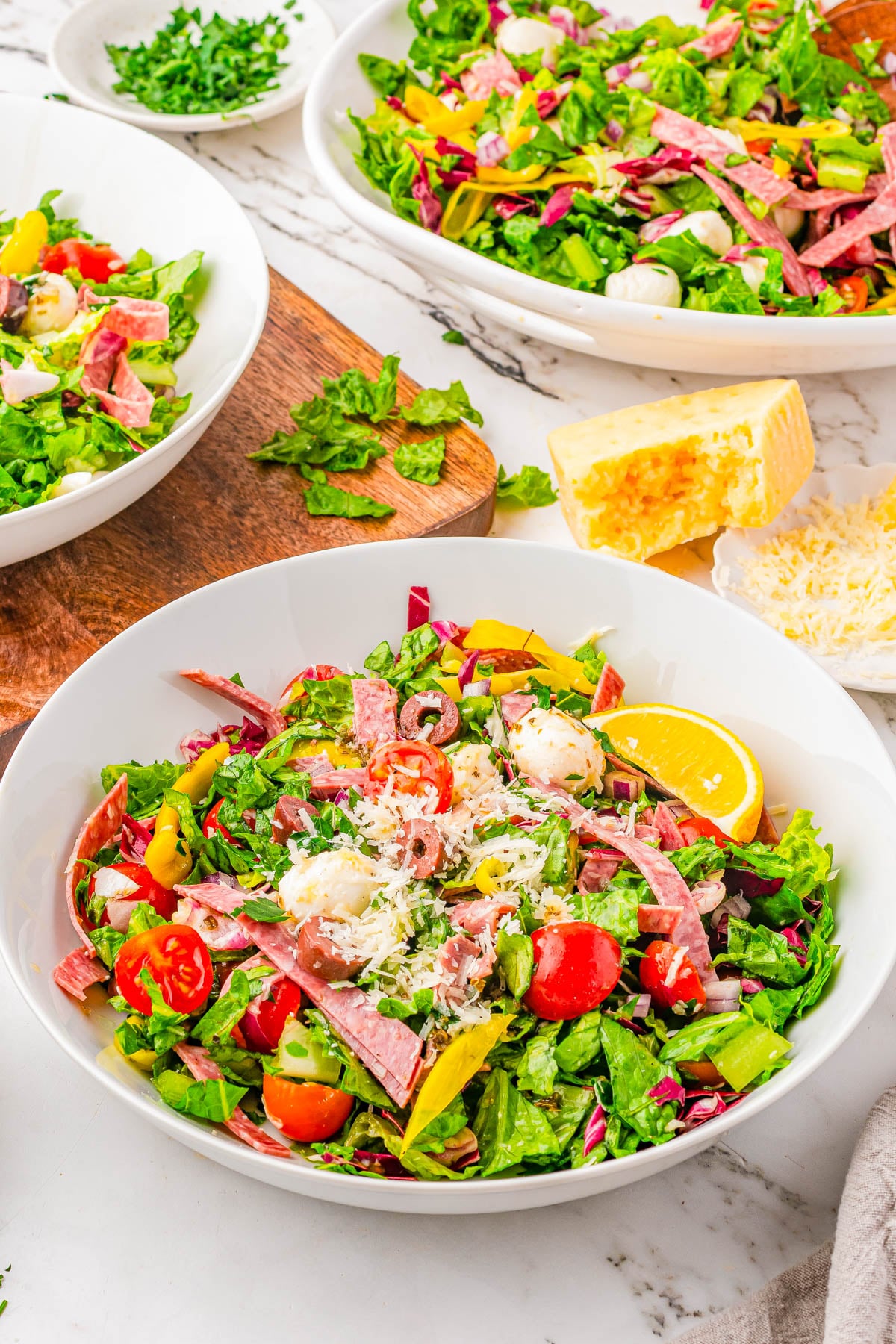A bowl of chopped salad with lettuce, tomatoes, salami, cheese, banana peppers, and a lemon wedge, with more salad and cheese in the background.