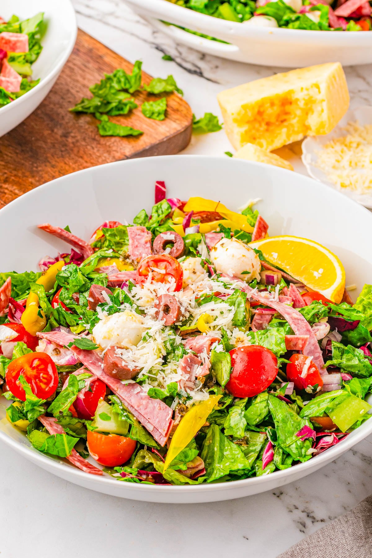 A bowl of chopped salad with lettuce, cherry tomatoes, sliced meats, pepperoncini, shredded cheese, and a lemon wedge on a marble table.