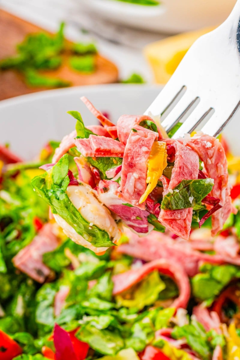 A fork holds a bite of salad with lettuce, sliced deli meat, cheese, and peppers over a bowl of mixed salad ingredients.