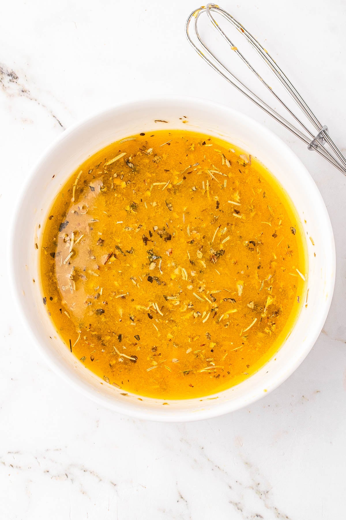 A white bowl filled with a yellow, herb-speckled vinaigrette next to a metal whisk on a white marble surface.
