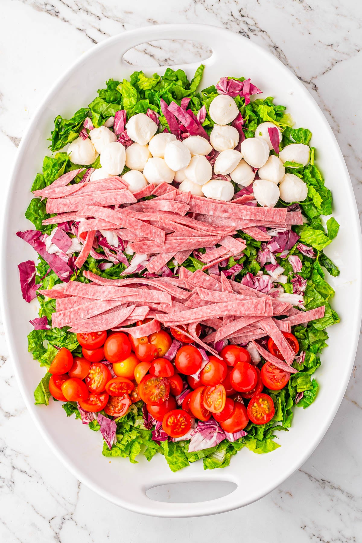 Oval platter with chopped lettuce, halved cherry tomatoes, sliced salami, mozzarella balls, and radicchio arranged in colorful rows on a marble surface.