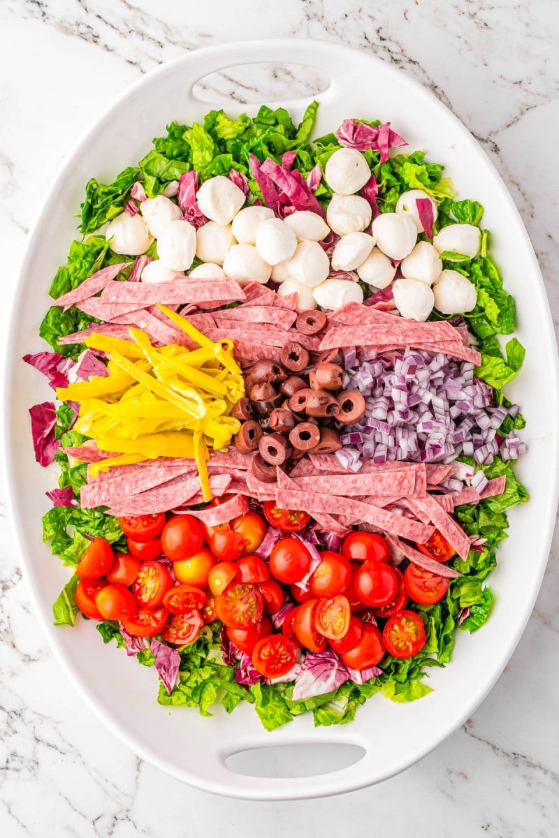 Oval platter with chopped lettuce, mozzarella balls, salami slices, yellow peppers, red onion, black olives, cherry tomatoes, and radicchio, arranged in neat rows on a marble surface.