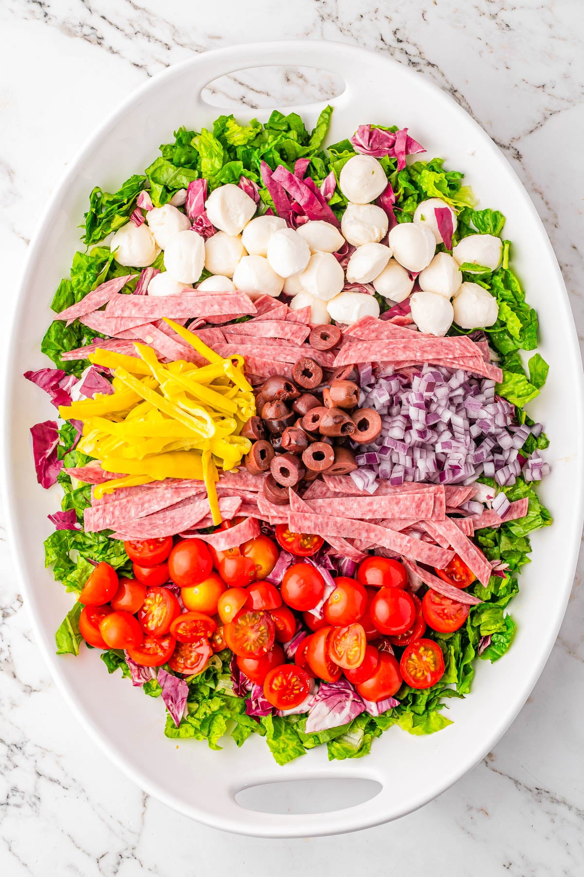 Oval platter with chopped lettuce, mozzarella balls, salami slices, yellow peppers, red onion, black olives, cherry tomatoes, and radicchio, arranged in neat rows on a marble surface.