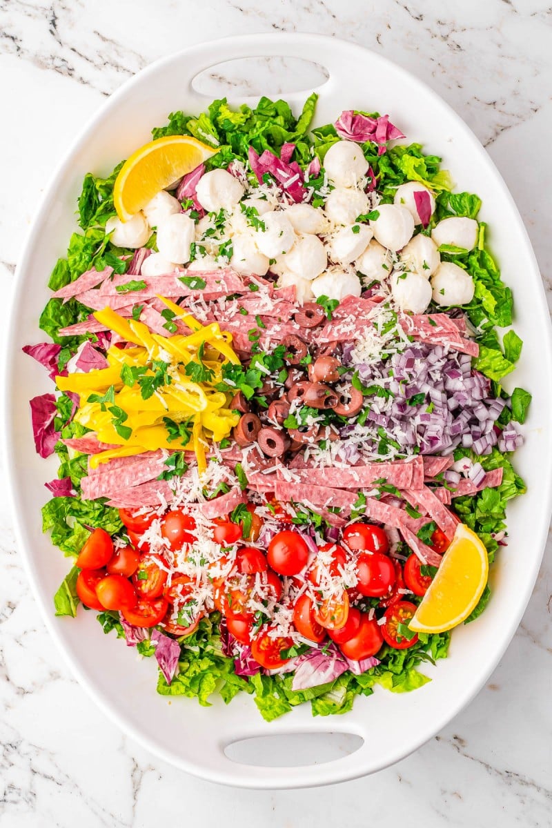 Oval platter with chopped lettuce, cherry tomatoes, salami strips, mozzarella balls, pepperoncini, olives, red onion, grated cheese, and lemon wedges, arranged in neat sections on a marble surface.
