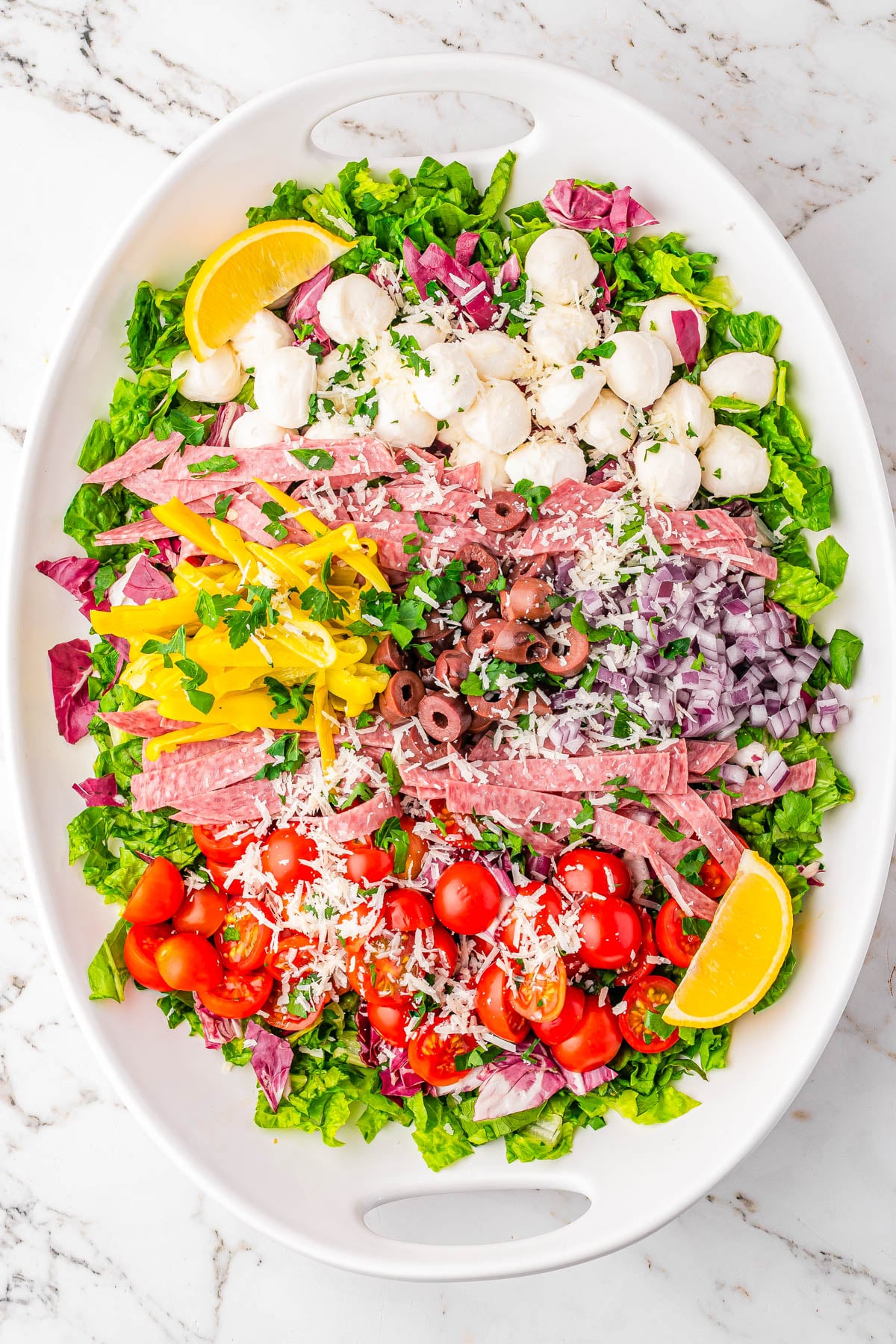Oval platter with chopped lettuce, cherry tomatoes, salami strips, mozzarella balls, pepperoncini, olives, red onion, grated cheese, and lemon wedges, arranged in neat sections on a marble surface.