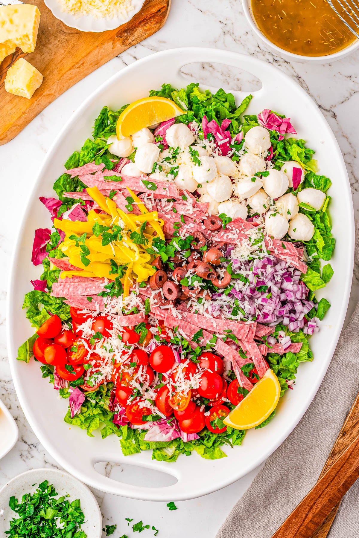 Oval platter with chopped lettuce, cherry tomatoes, red onion, sliced salami, mozzarella balls, pepperoncini, olives, shredded cheese, and lemon wedges, arranged in sections.
