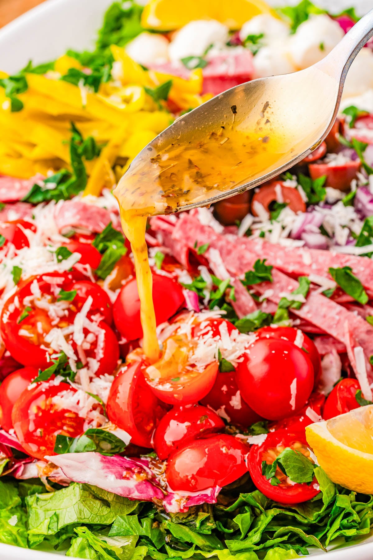 A spoon pours vinaigrette dressing over a salad with cherry tomatoes, sliced salami, shredded cheese, greens, and lemon wedges.