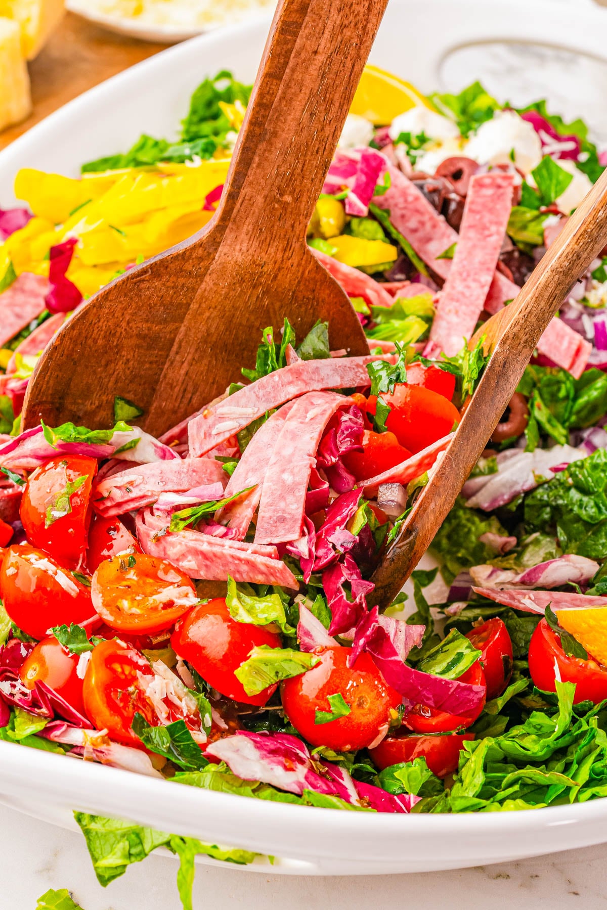 A close-up of a chopped salad with lettuce, cherry tomatoes, salami strips, banana peppers, olives, and wooden salad tongs in a white bowl.