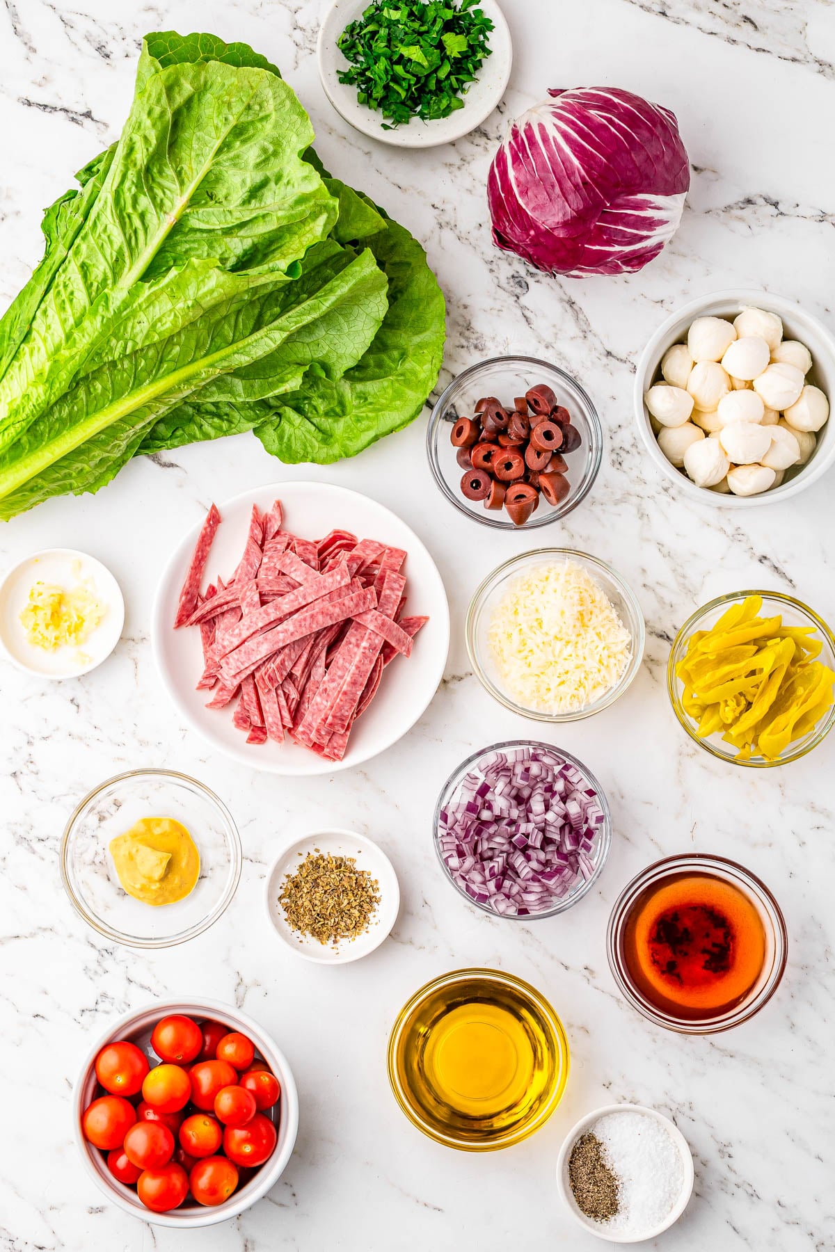 Various salad ingredients, including lettuce, radicchio, cherry tomatoes, sliced salami, onions, olives, mozzarella balls, cheese, dressing, oil, seasonings, and herbs on a marble surface.