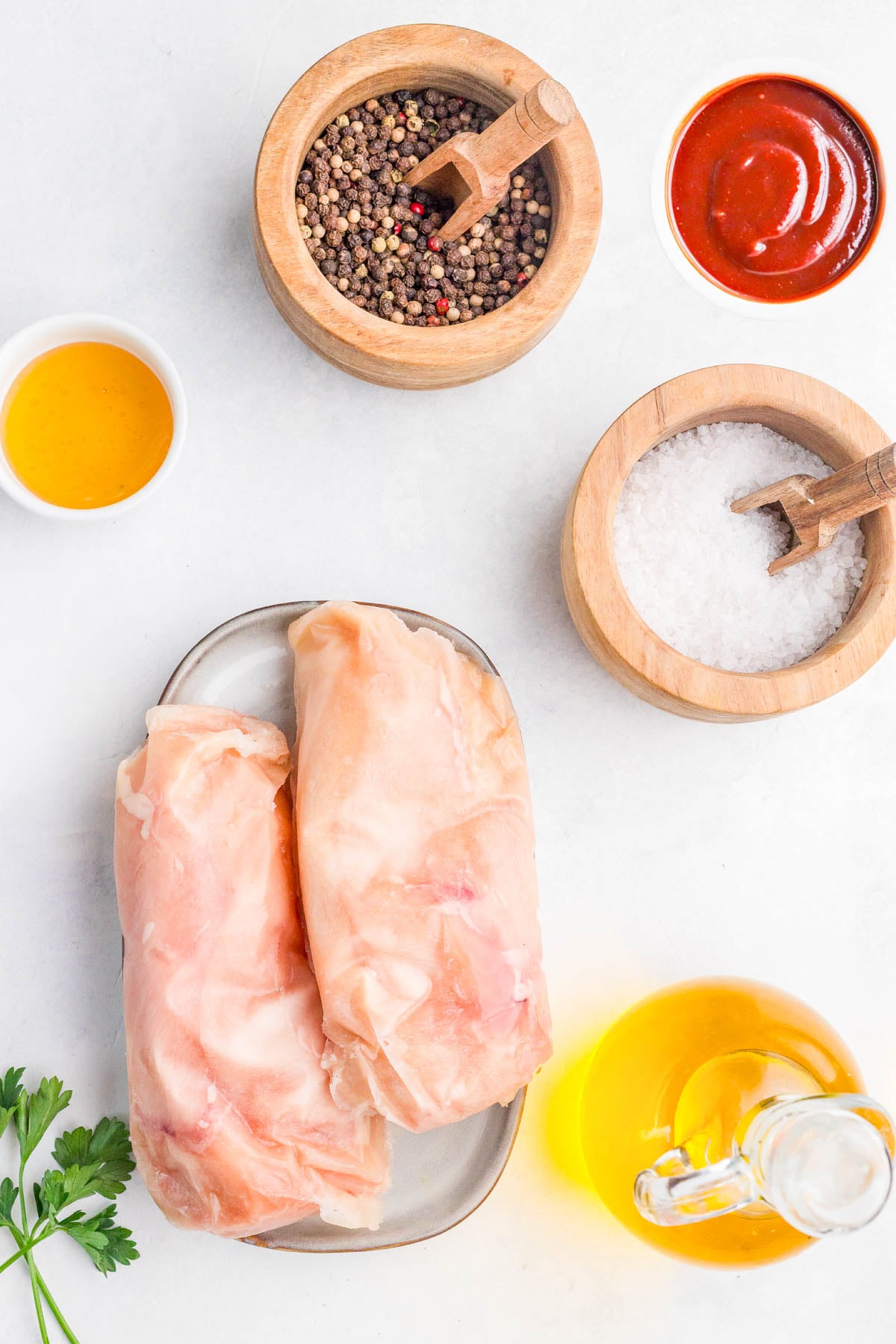 Raw chicken breasts on a plate, surrounded by black peppercorns, coarse salt, barbecue sauce, oil, and honey on a white surface.
