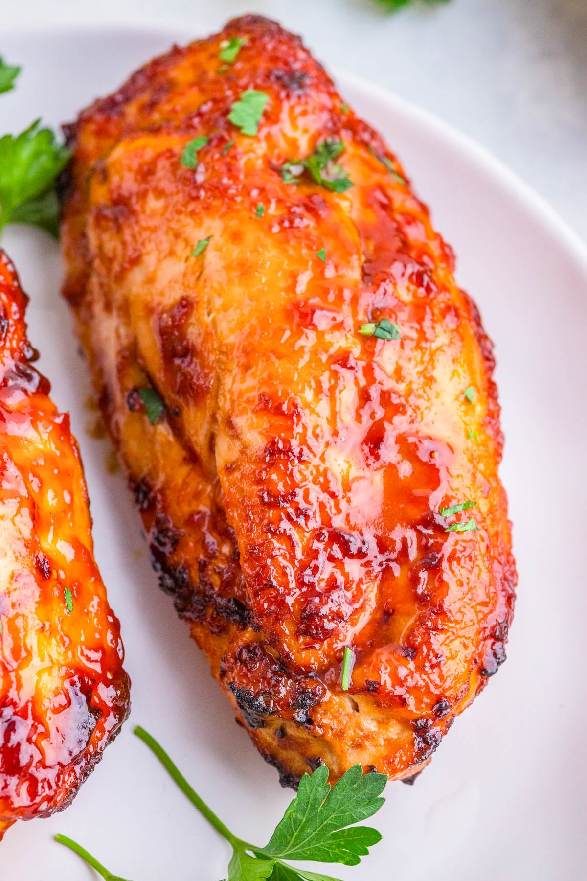 Close-up of a glazed, cooked chicken breast garnished with chopped parsley on a white plate.