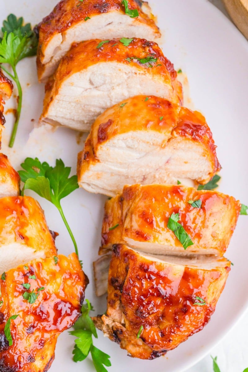 Sliced grilled chicken breast topped with a glossy orange sauce, garnished with chopped parsley on a white plate.