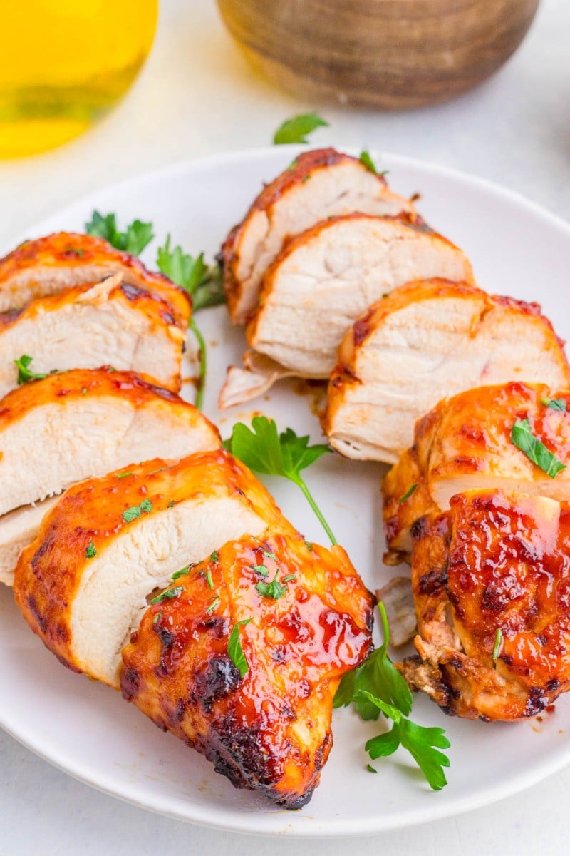 Sliced grilled chicken breasts with a glazed sauce and parsley garnish are arranged on a white plate.
