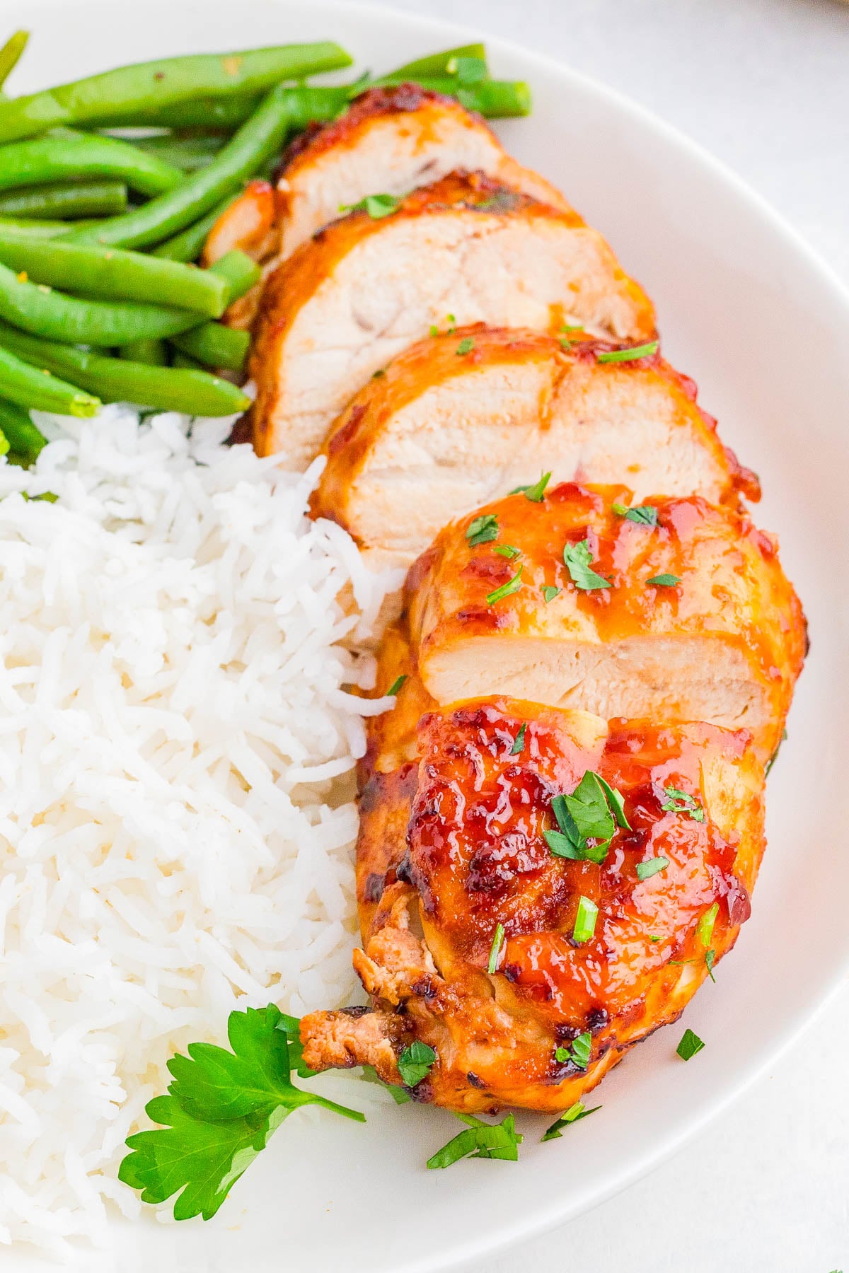 Sliced grilled chicken breast with sauce, served with white rice and green beans on a white plate.