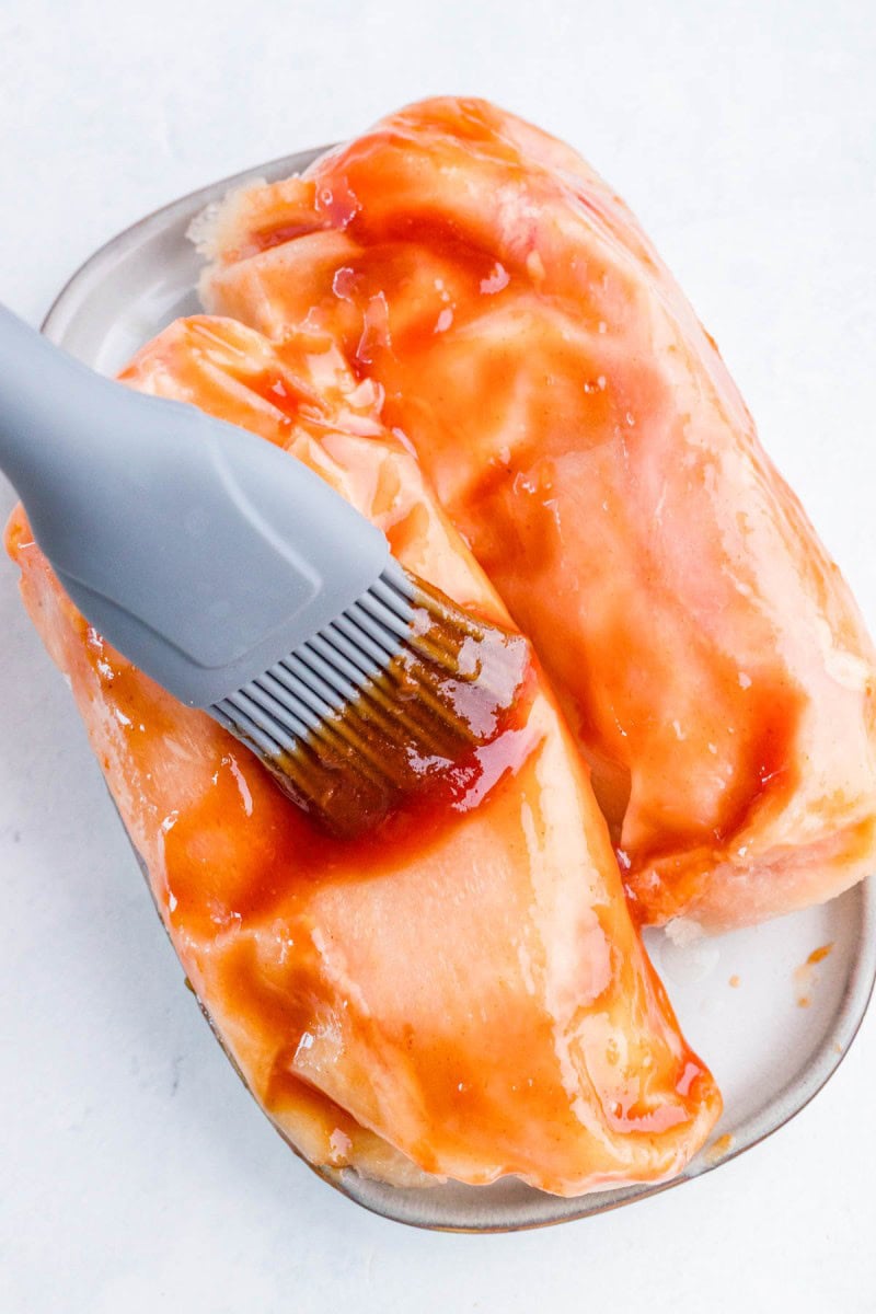 A silicone brush spreads reddish sauce onto raw chicken breasts on a small oval plate.