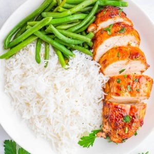 A white plate with sliced glazed chicken breast, steamed green beans, and a serving of white rice, garnished with parsley.