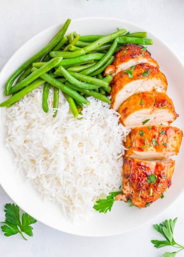 A white plate with sliced glazed chicken breast, steamed green beans, and a serving of white rice, garnished with parsley.