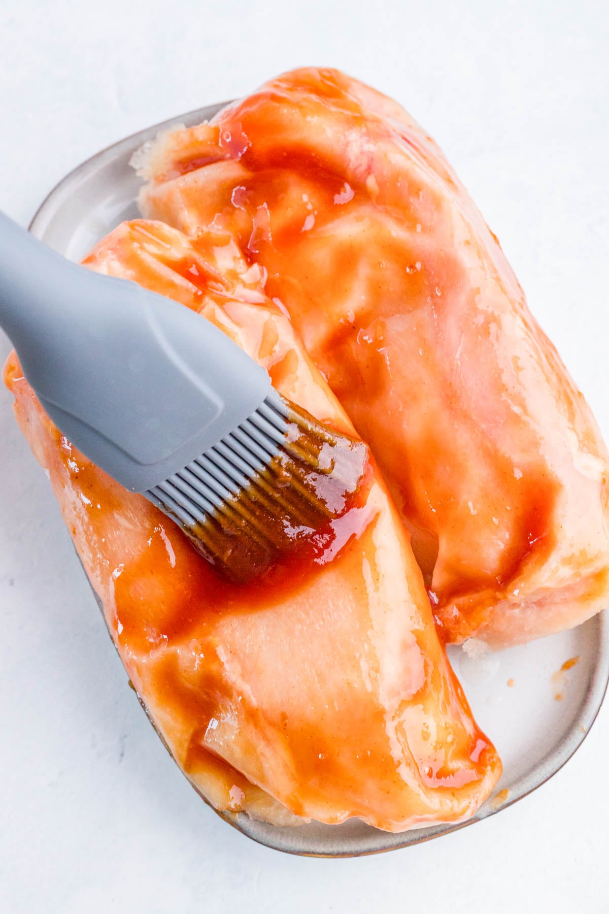 A silicone brush spreads reddish sauce onto raw chicken breasts on a small oval plate.