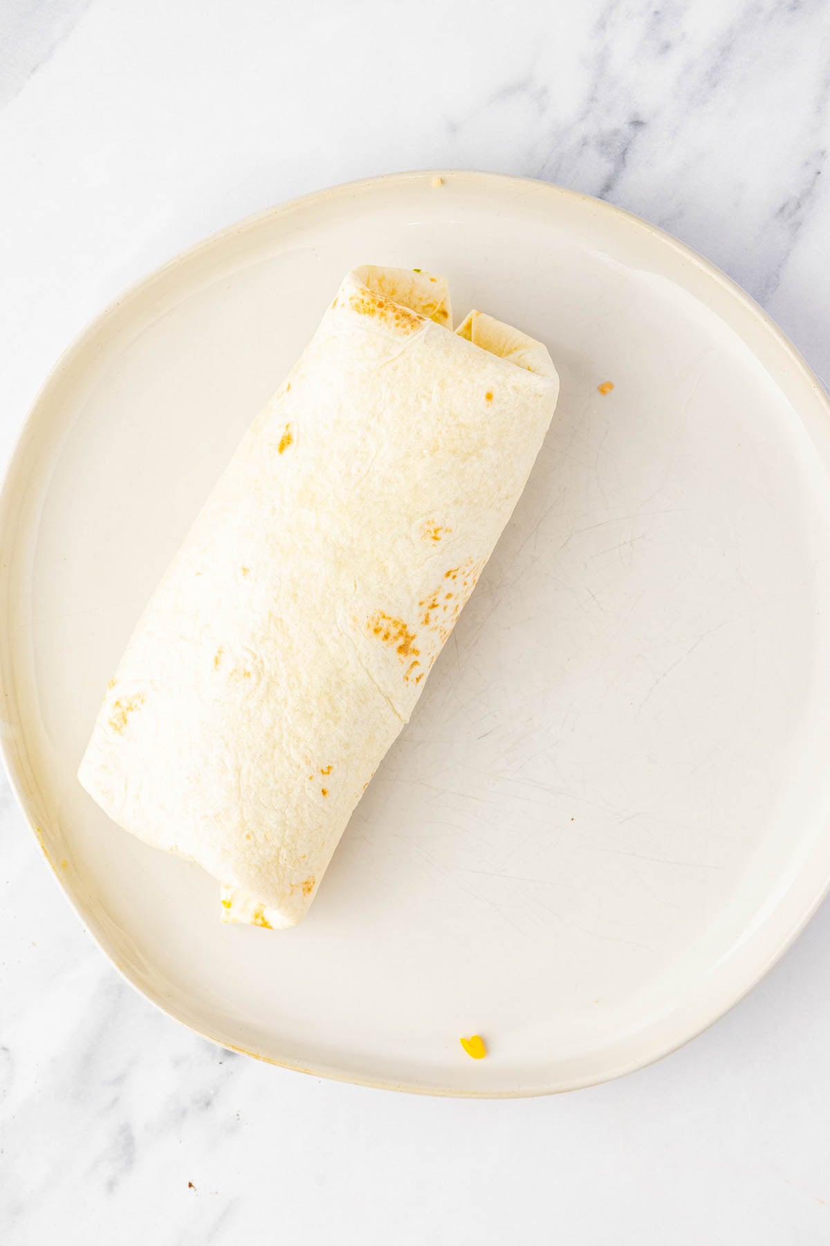 A wrapped burrito is placed on a round white plate set on a marble surface.