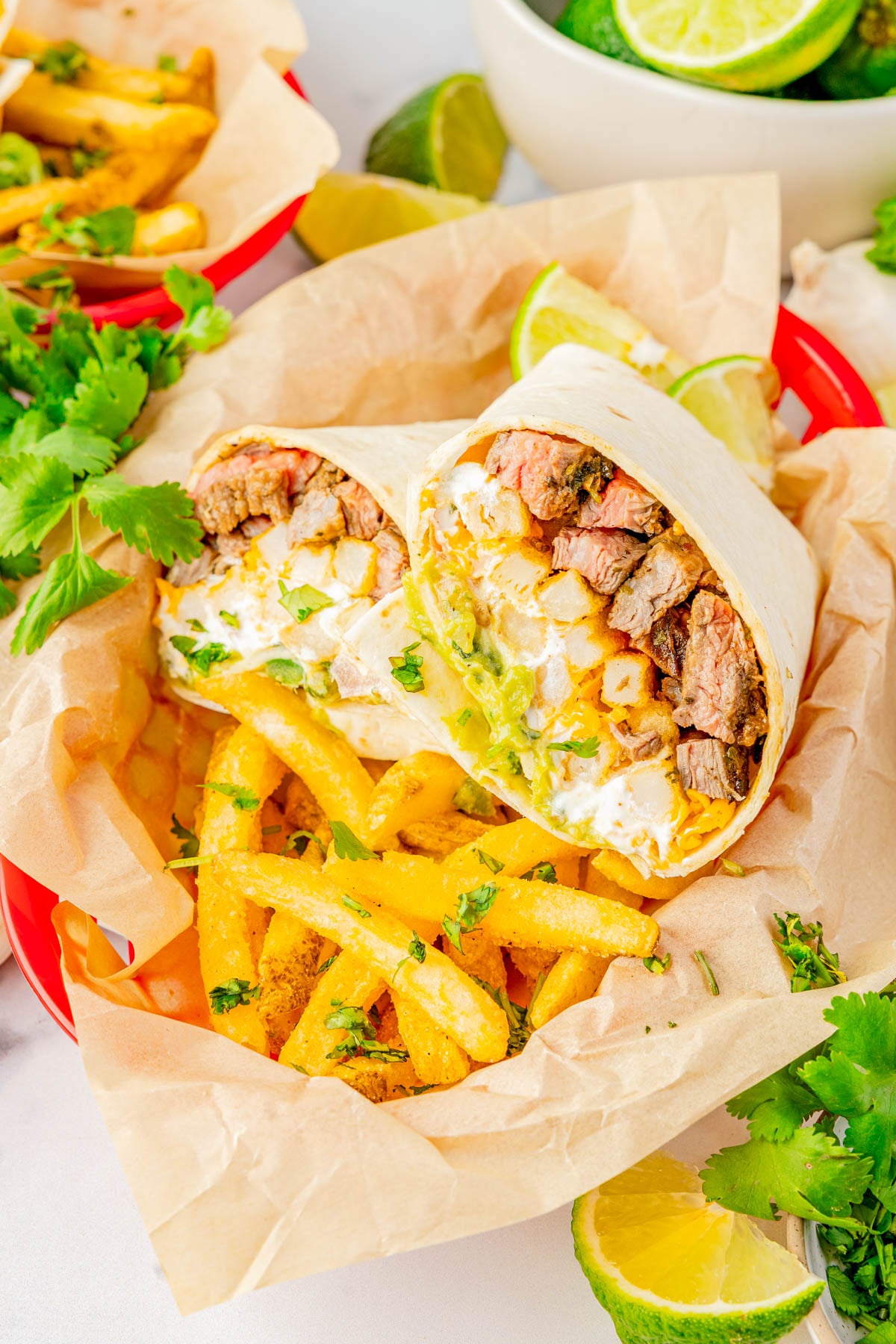 A beef and potato burrito cut in half served with seasoned fries in a red basket, garnished with lime wedges and fresh cilantro.