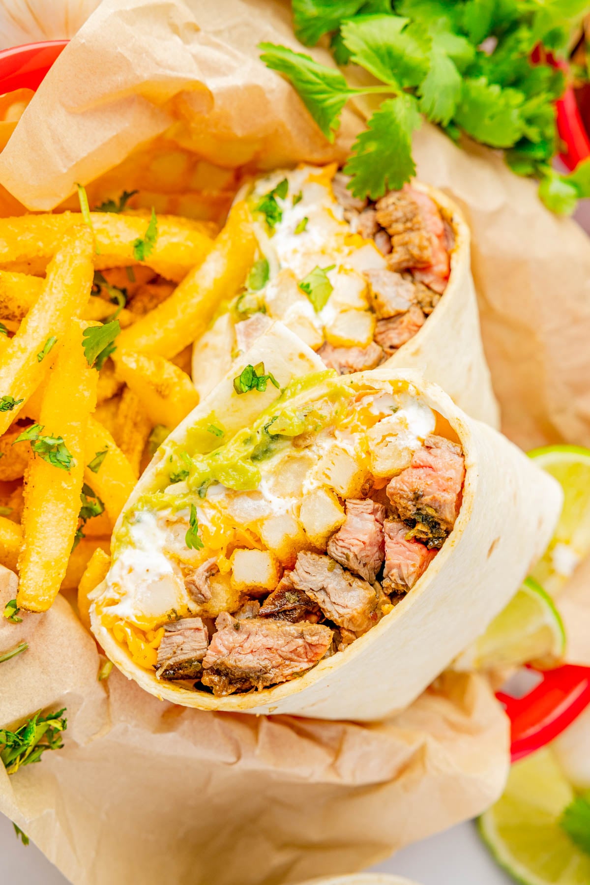 A steak burrito cut in half with fries on the side, garnished with cilantro, served in a red basket lined with parchment paper.