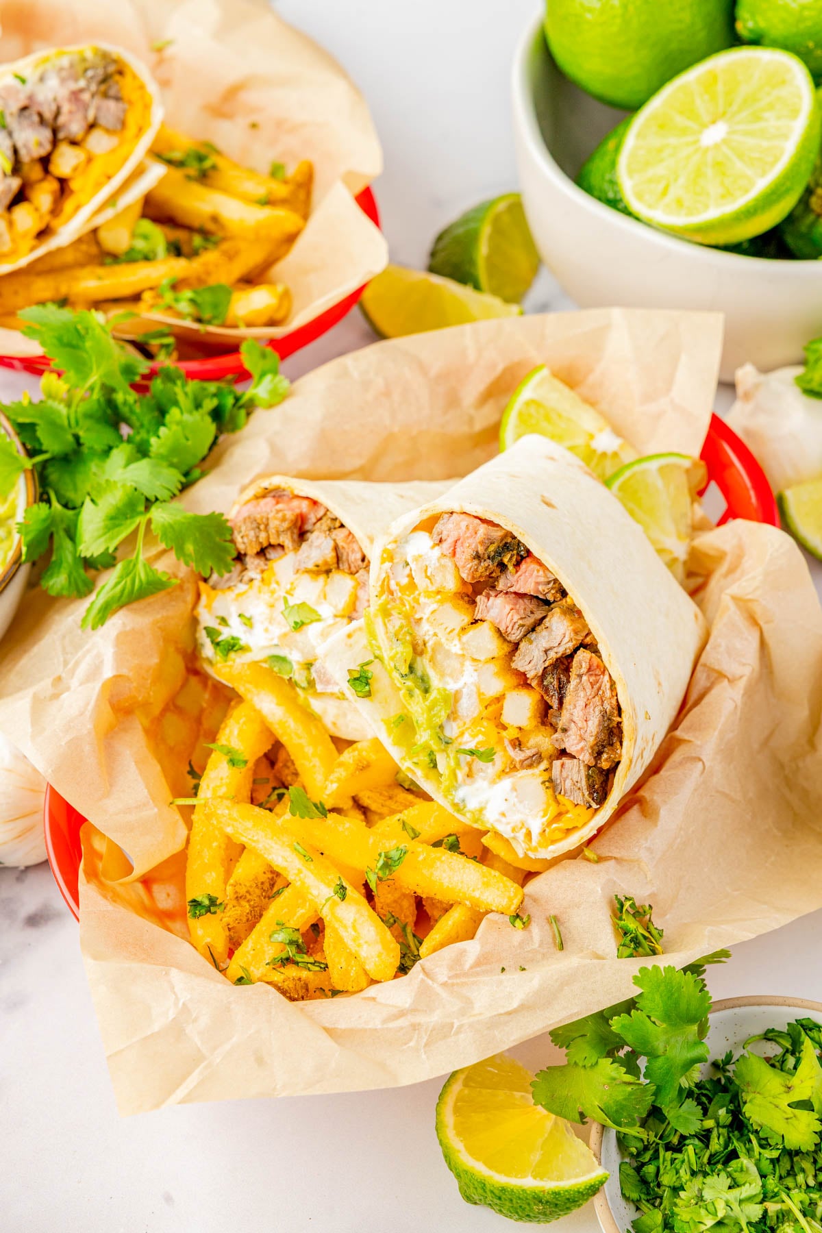 A burrito cut in half filled with steak, French fries, cheese, and cilantro, served on parchment paper with lime wedges and fresh cilantro.