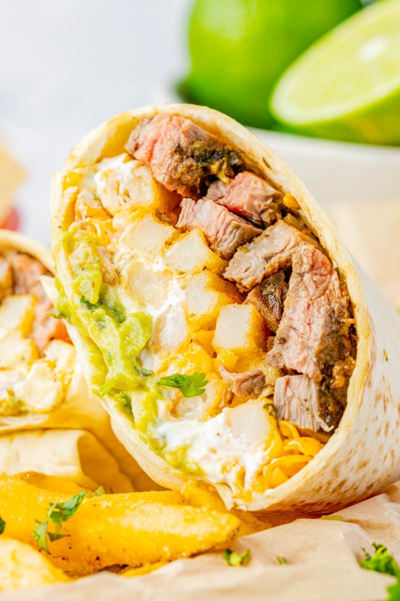 A close-up of a sliced steak burrito filled with steak pieces, cheese, fries, guacamole, and sour cream, with lime and fries in the background.