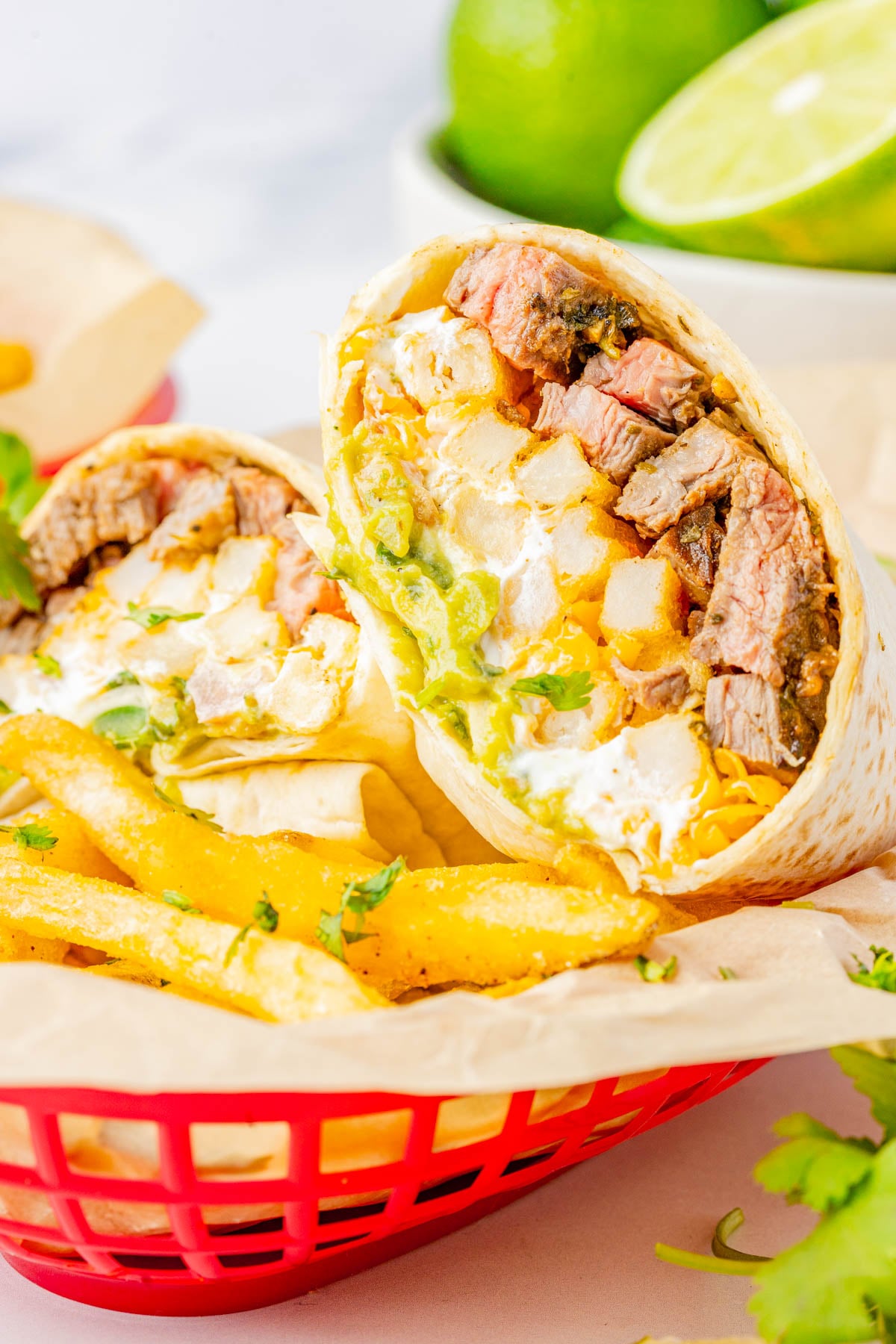 A burrito filled with steak, rice, cheese, sour cream, and guacamole, cut in half and served with French fries in a red basket.