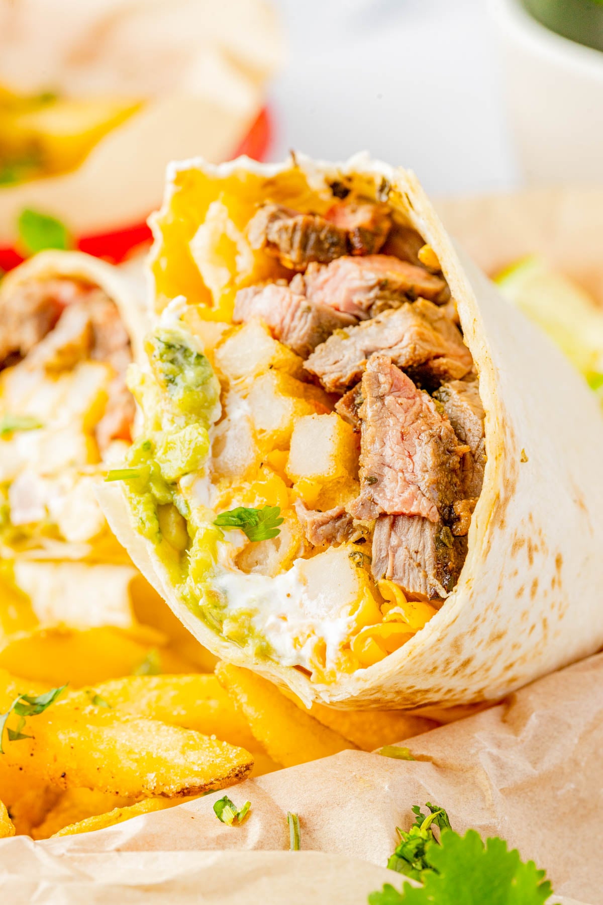 A close-up of a burrito cut in half, filled with grilled steak, cheese, guacamole, sour cream, and potatoes, served with French fries on parchment paper.