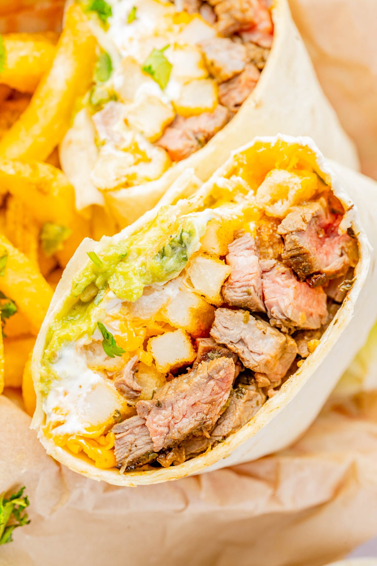 A close-up of a burrito cut in half, filled with steak, cheese, fries, guacamole, and sour cream, with curly fries on the side.