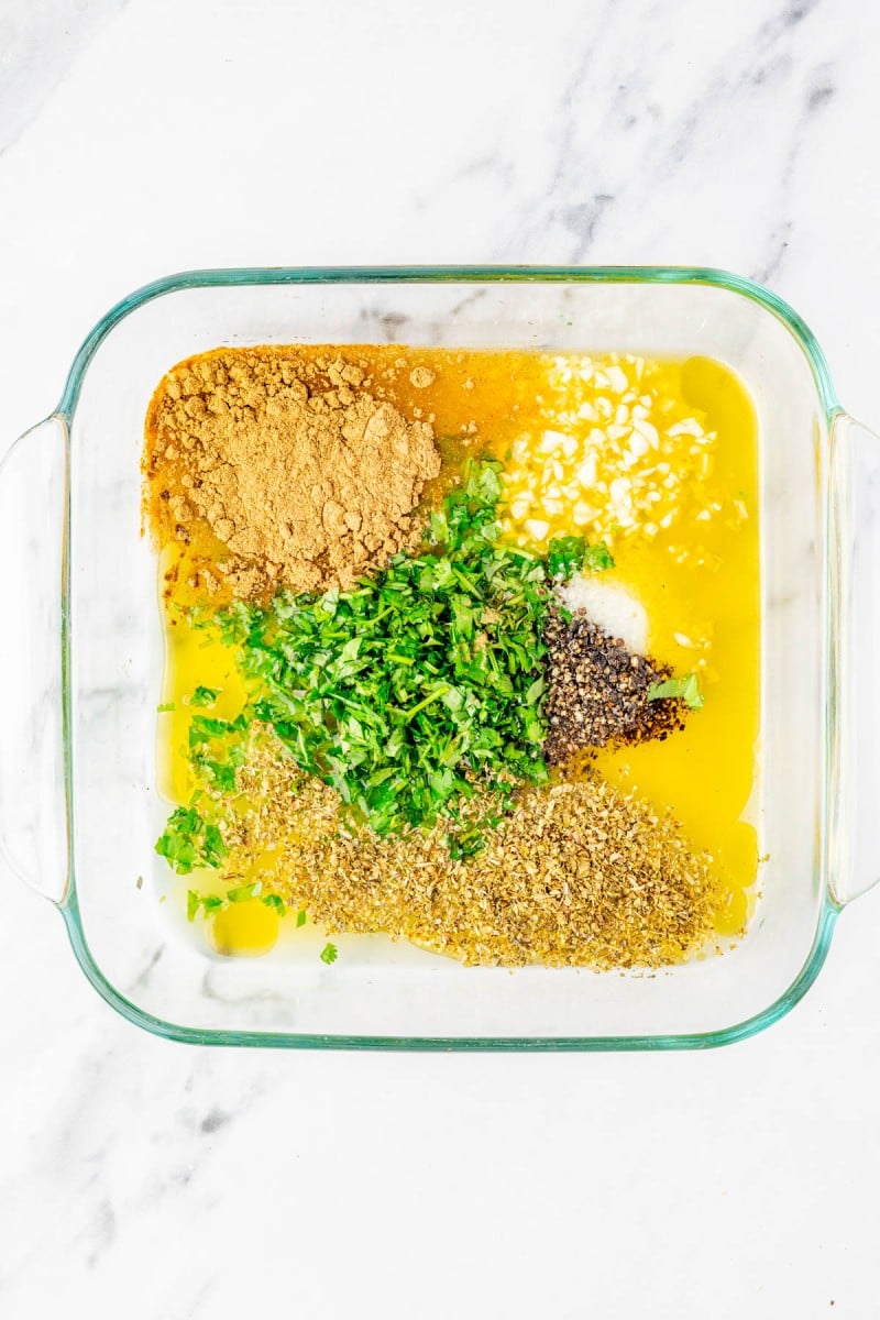A glass dish containing melted butter, chopped herbs, ground spices, minced garlic, salt, and pepper, all separated and unmixed on a white marble surface.