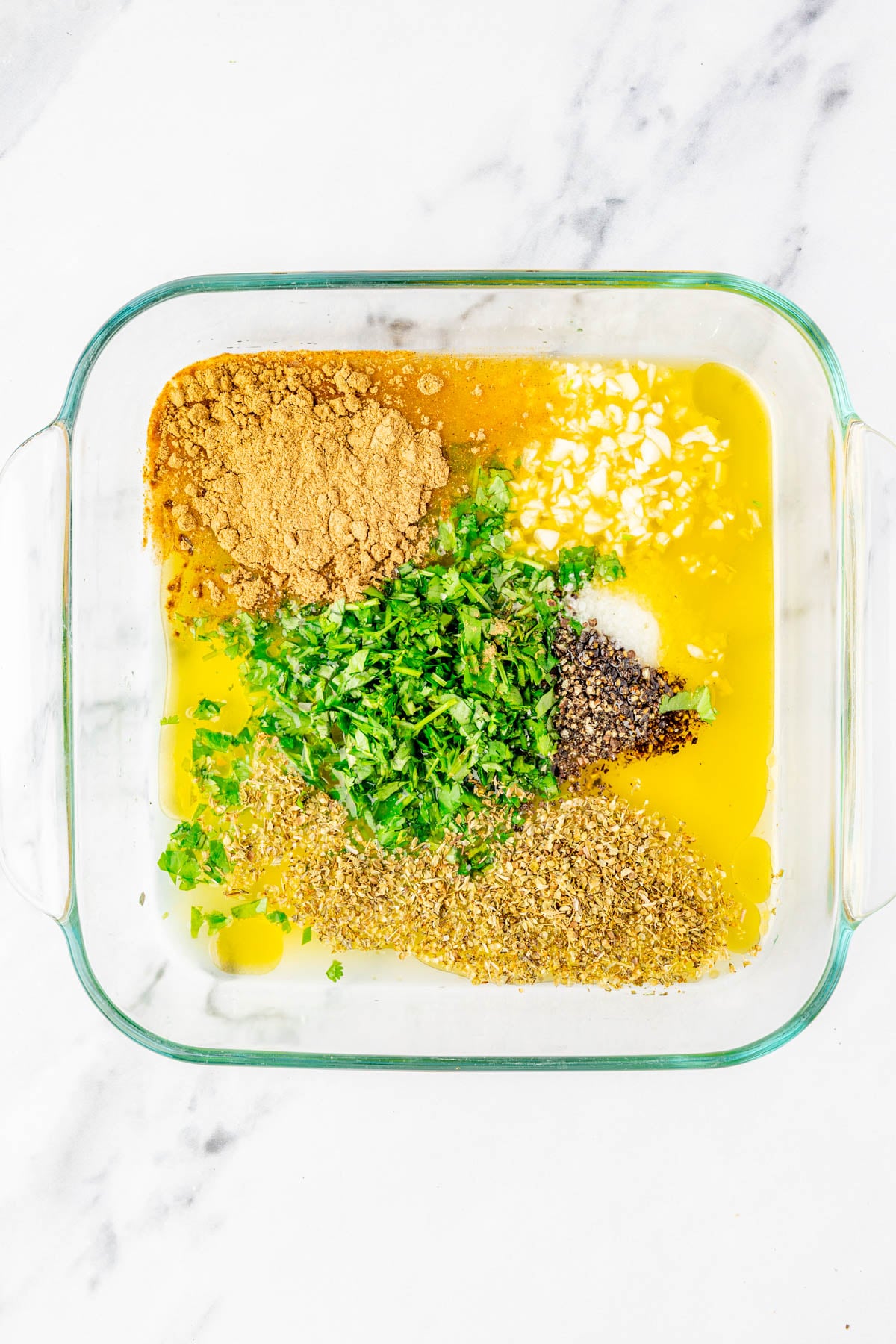 A glass dish containing melted butter, chopped herbs, ground spices, minced garlic, salt, and pepper, all separated and unmixed on a white marble surface.