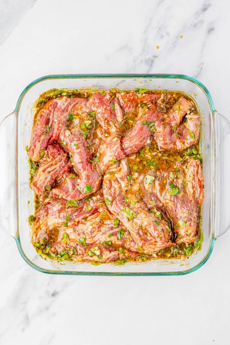 Raw steak marinating in a glass dish with herbs and spices on a white marble surface.