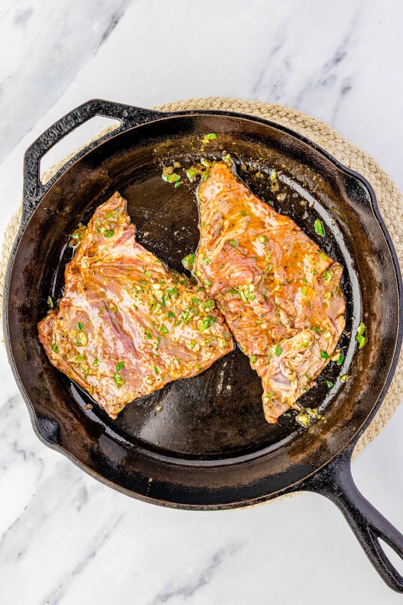 Two marinated pieces of meat with chopped herbs are cooking in a black cast iron skillet on a marble surface.