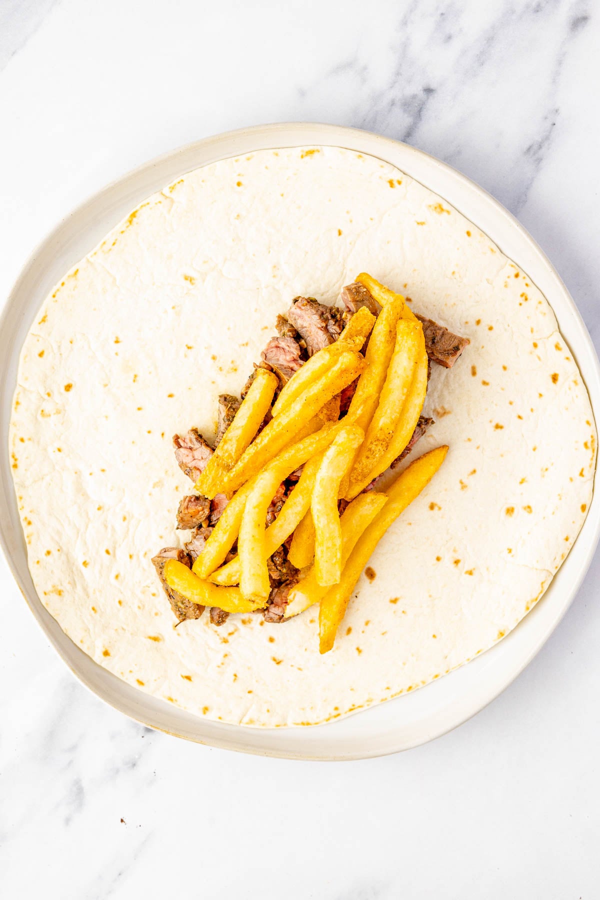 A flour tortilla on a plate with steak pieces and French fries in the center, ready to be wrapped.