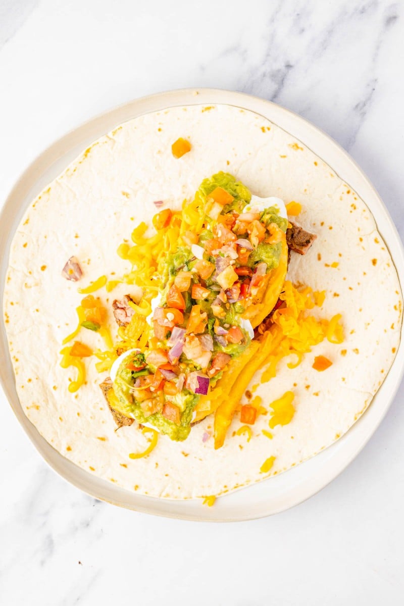 A flour tortilla topped with shredded cheese, lettuce, diced tomatoes, onions, and chopped vegetables on a white plate.