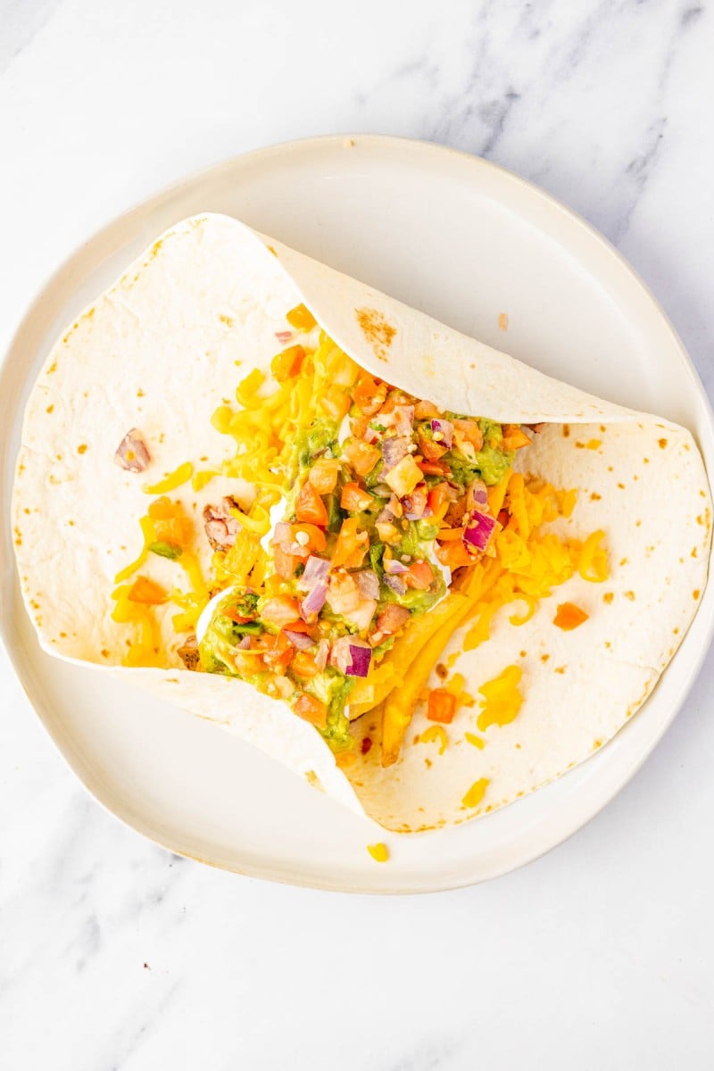 A flour tortilla on a white plate filled with shredded cheddar cheese, guacamole, and fresh salsa, ready to be wrapped.