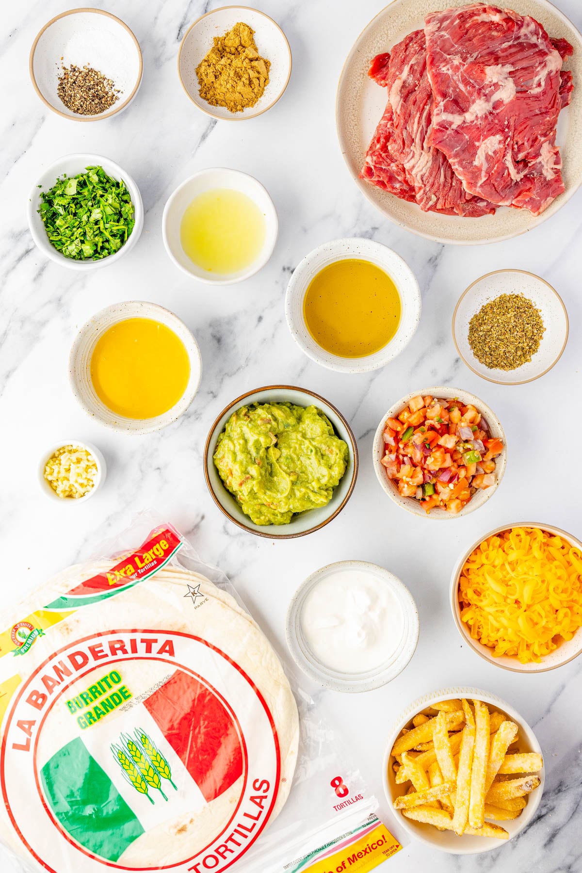Ingredients for burritos arranged on a white surface, including tortillas, sliced beef, shredded cheese, guacamole, pico de gallo, sour cream, chopped herbs, seasonings, and sauces.