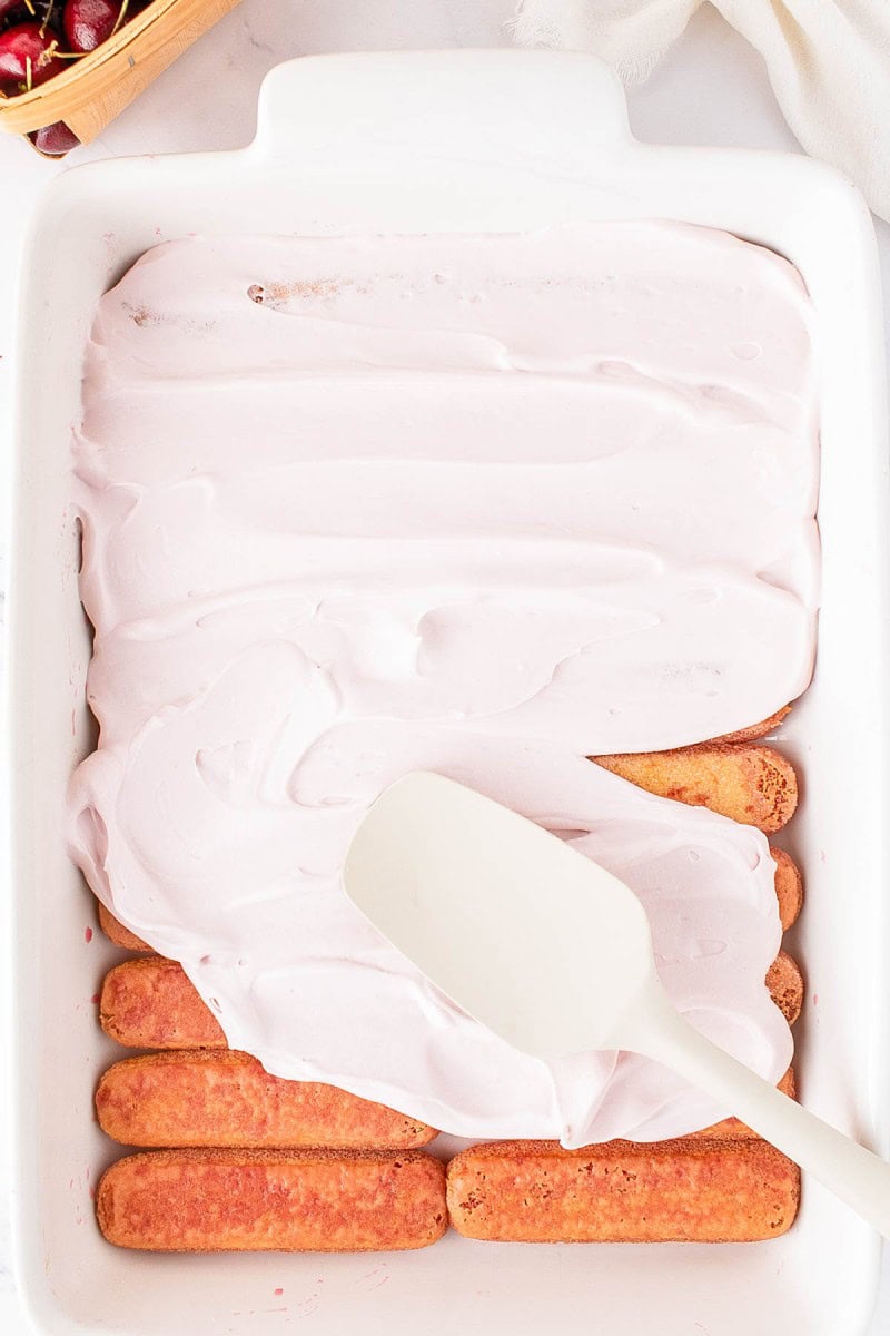 A white spatula spreads pink cream over a layer of ladyfinger cookies in a white baking dish.