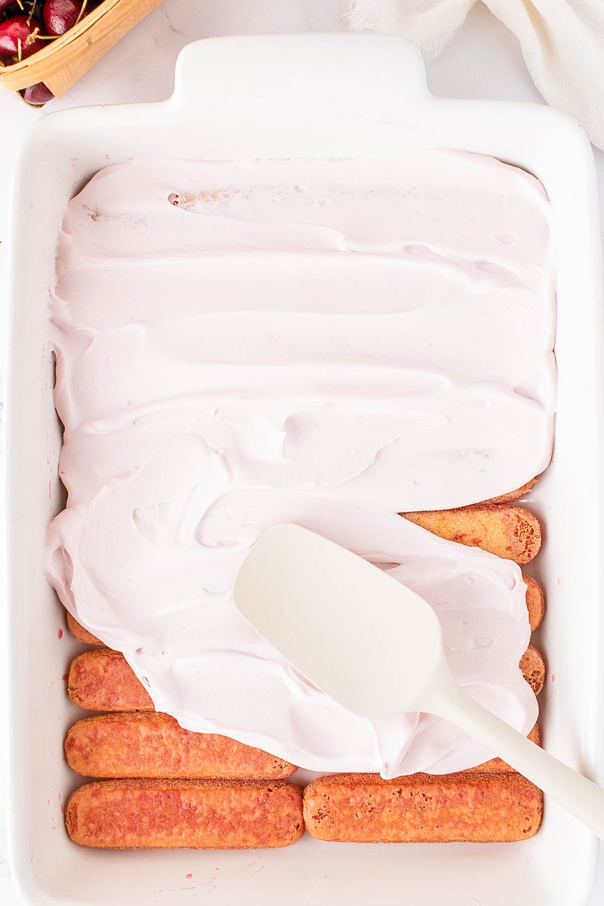 A white spatula spreads pink cream over a layer of ladyfinger cookies in a white baking dish.