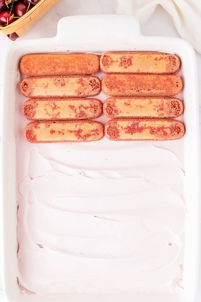 A white dish with a layer of pink cream and a row of seven ladyfingers on top, next to a basket of cherries in the corner.