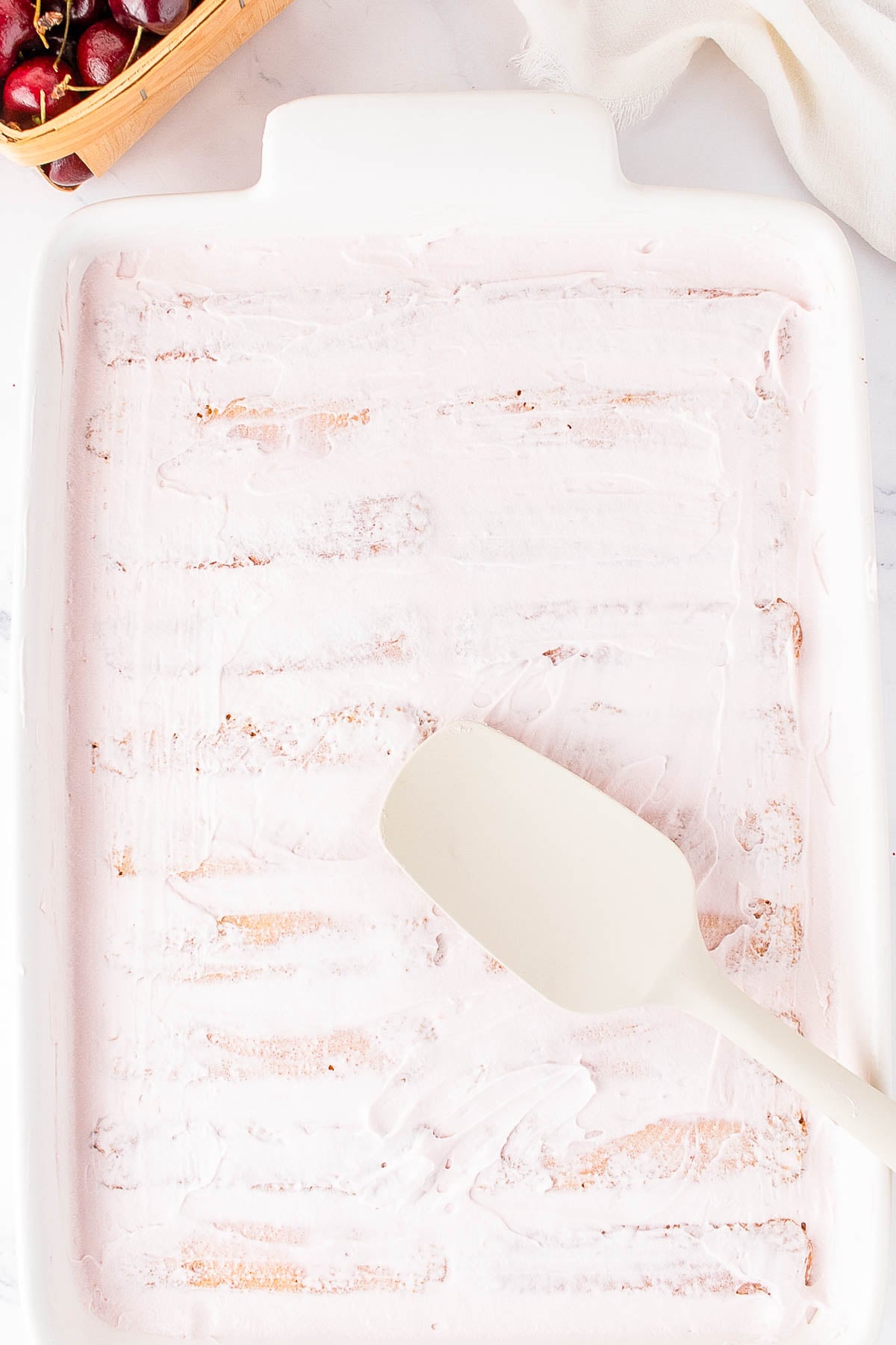A white spatula rests in a rectangular white dish with a thin, even layer of pink frosting spread across the bottom.