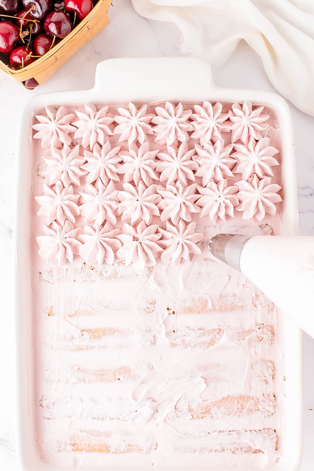 A hand pipes pink whipped topping in star shapes onto a partially decorated dessert in a white rectangular baking dish; cherries sit in a basket nearby.