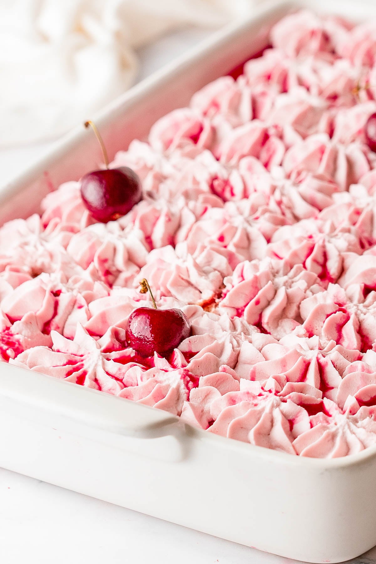 A white rectangular dish filled with piped pink whipped topping and garnished with a few whole cherries.