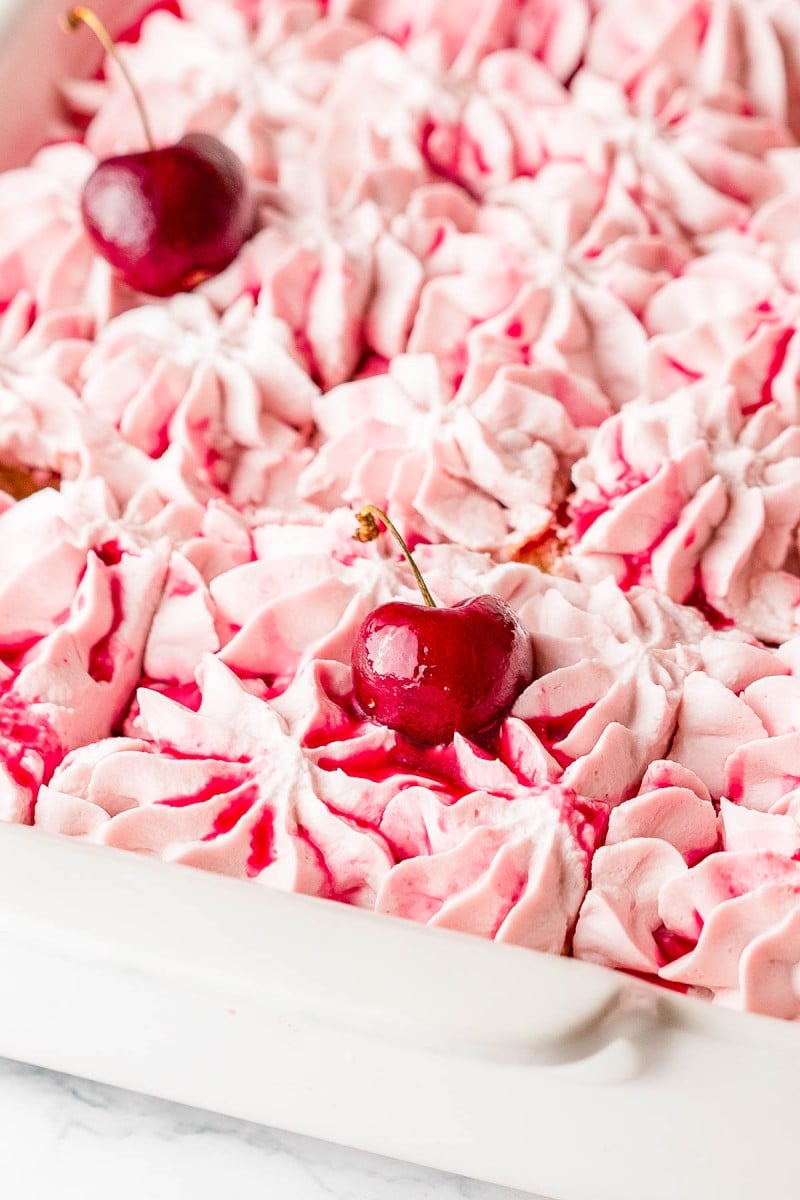 Close-up of pink whipped cream piped in swirls in a white dish, with two cherries and red syrup drizzled on top.