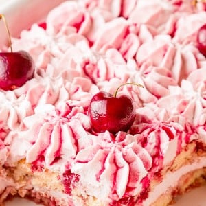 A close-up of a cherry dessert topped with pink whipped cream and whole cherries, with a portion removed to show the layered interior.