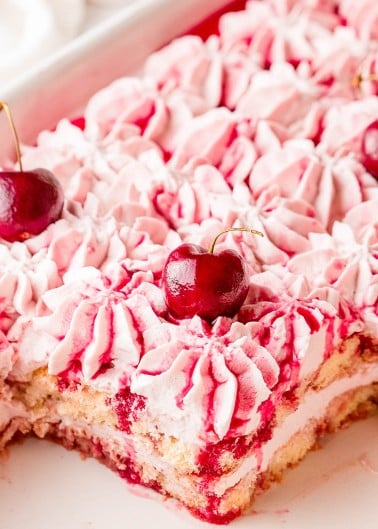 A close-up of a cherry dessert topped with pink whipped cream and whole cherries, with a portion removed to show the layered interior.