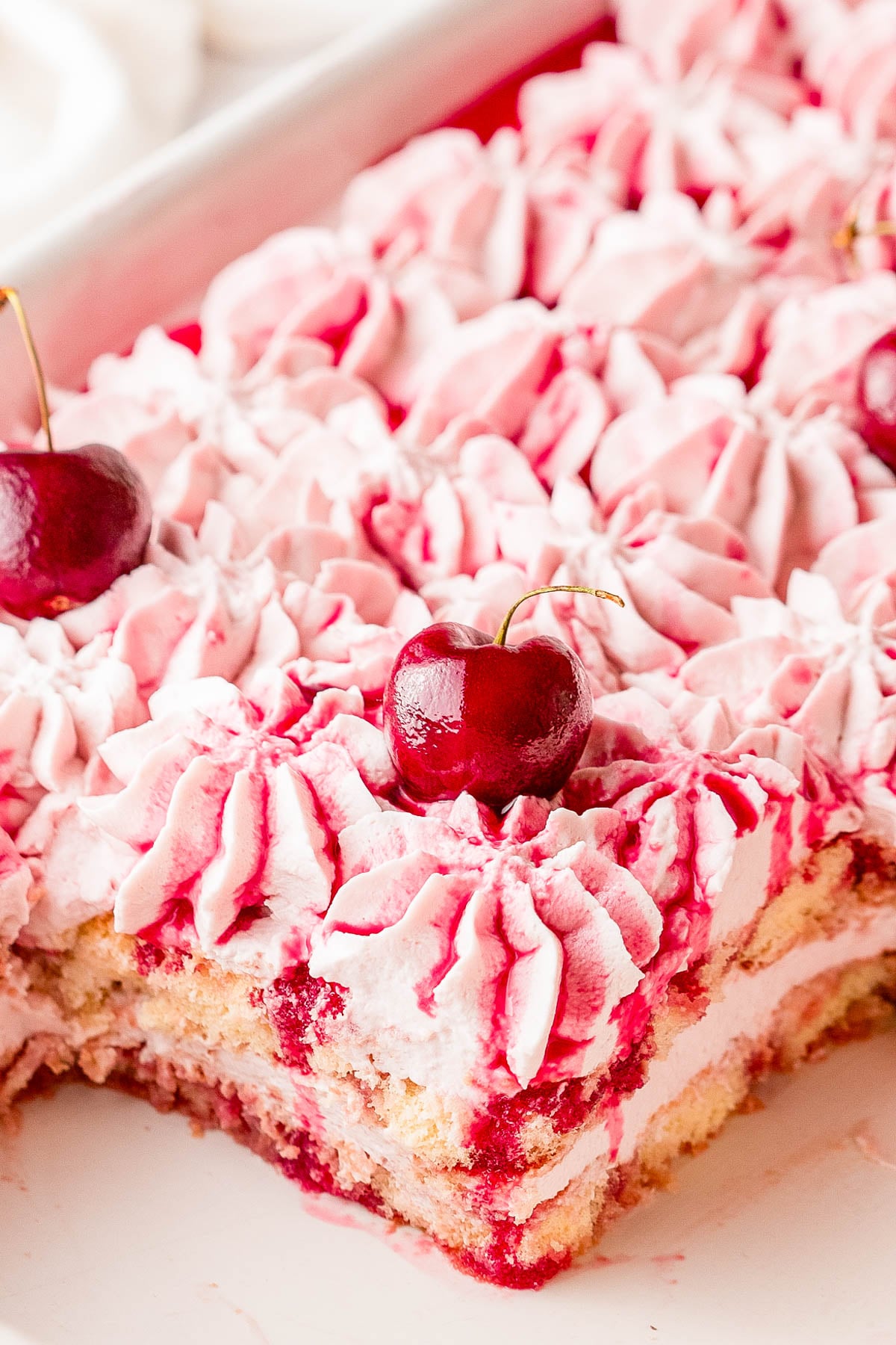 A close-up of a cherry dessert topped with pink whipped cream and whole cherries, with a portion removed to show the layered interior.
