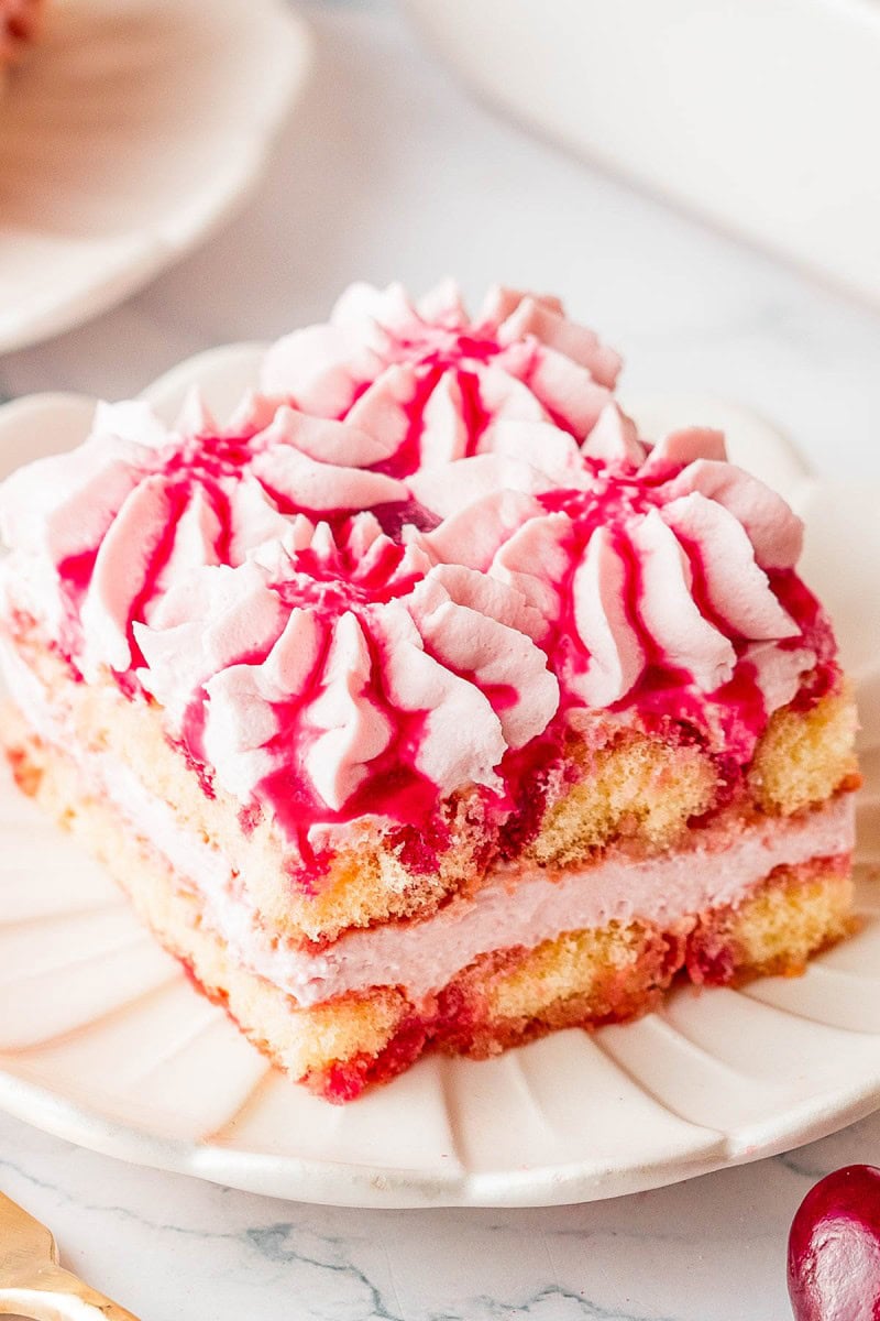 A square slice of layered sponge cake with pink frosting and decorative piped cream, served on a white plate.